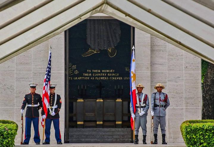 Members of the American and Philippine military stand with flags and weapons outside the chapel building.