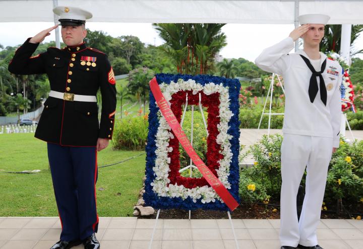 A marine and a sailor salute next to a floral wreath. 