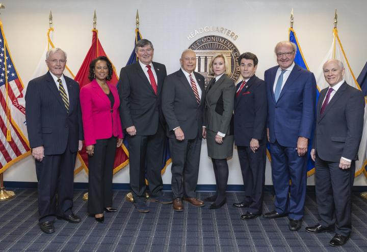 ABMC commissioners stand in front of ABMC seal, American flag and service flags. 