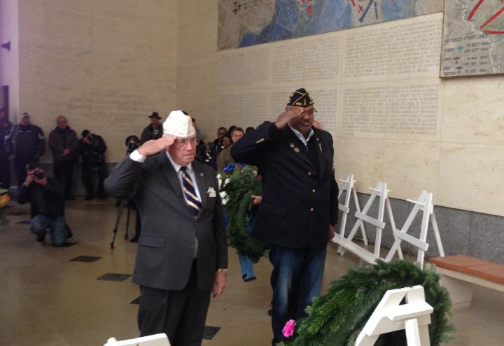 Two men stand and salute after laying floral wreaths.