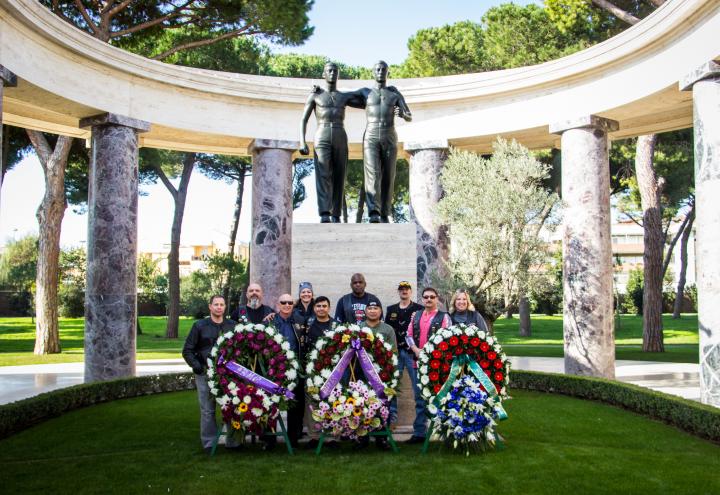 Men and women in motorcycle gear stand behind large floral wreaths after the ceremony. 