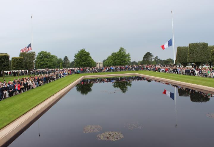 Attendees sit around the reflecting pool, with the American and French flag at half mast in the background. 