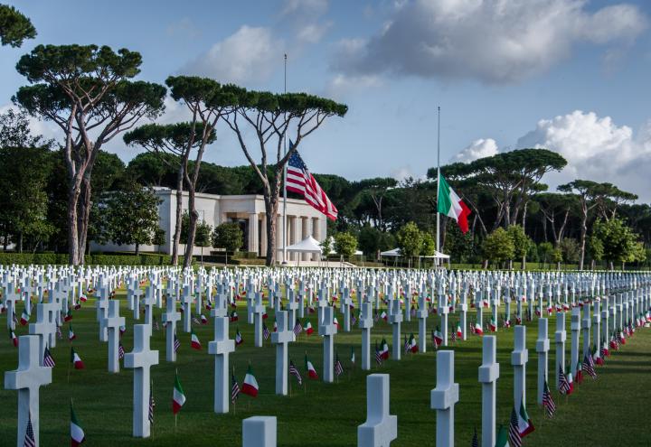 Rows of headstones with American and Italian flags cover the landscape in front of the memorial building.