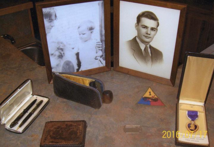 A picture frame,pipe, wallet, pens, a purple heart, a unit patch, and a dog tag are gathered on a countertop. 