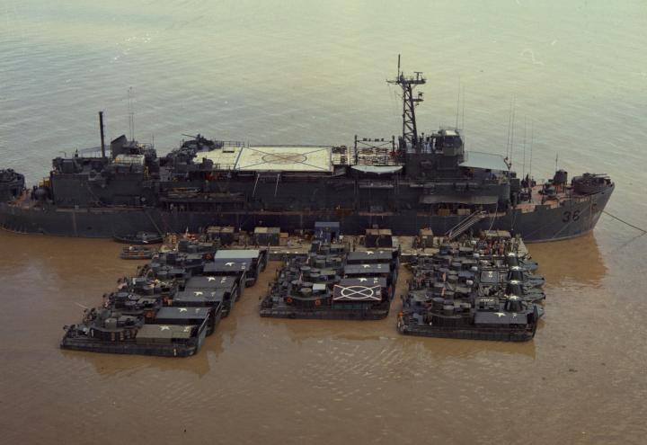 Large ship with floating pier and helipad next to grouping of 15 smaller vessels. 