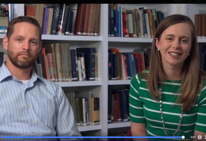 Man and woman sit in front of bookshelves. 