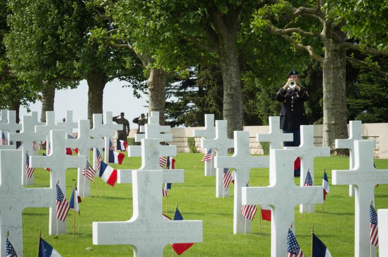 A bugler stands amidst headstones, each marked with an American and French flag. 